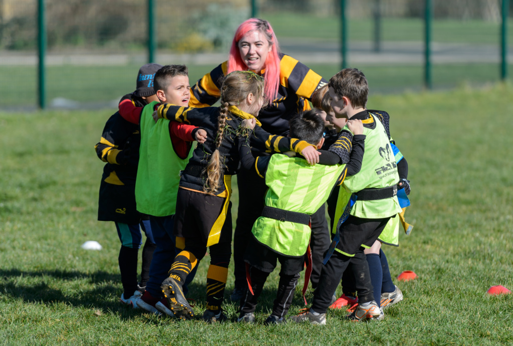 Alt Text: Adult with long pink hair coaching groups children playing rugby.